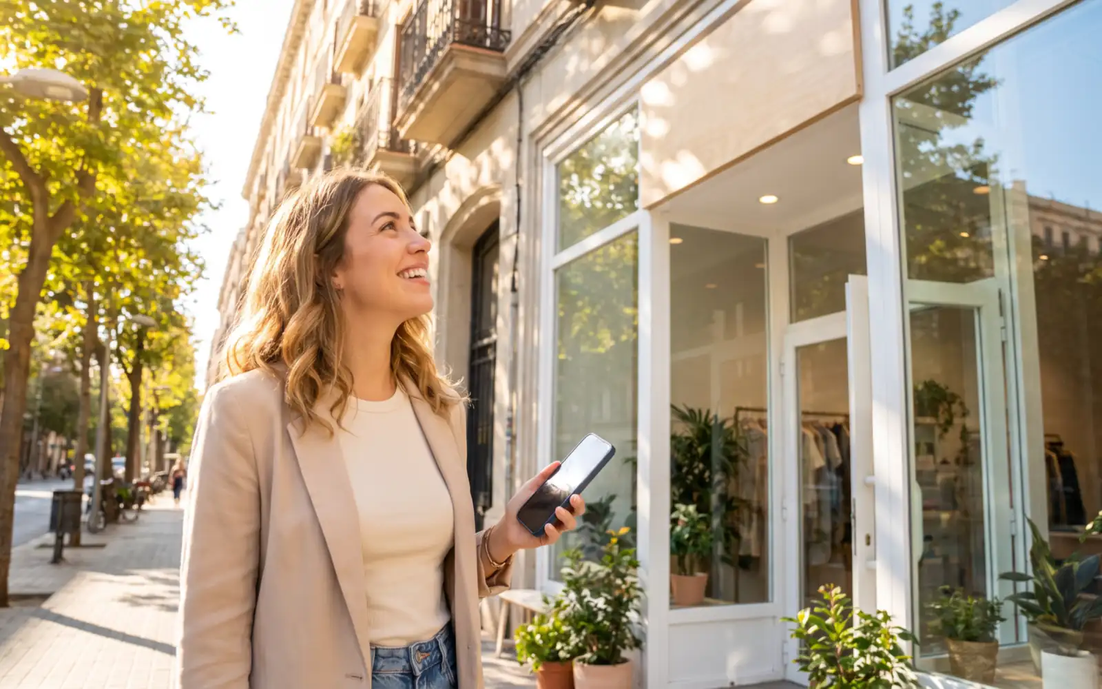A smiling woman in a blazer holding her phone on a sunny, tree-lined sidewalk outside a bright boutique storefront